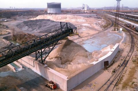 Tas de laitier de haut fourneau à l’usine sidérurgique de Fos-sur-Mer : laitier vitrifié  (en beige clair, cône au premier plan et tas en arrière-plan)  et cristallisé (en gris, au centre gauche)